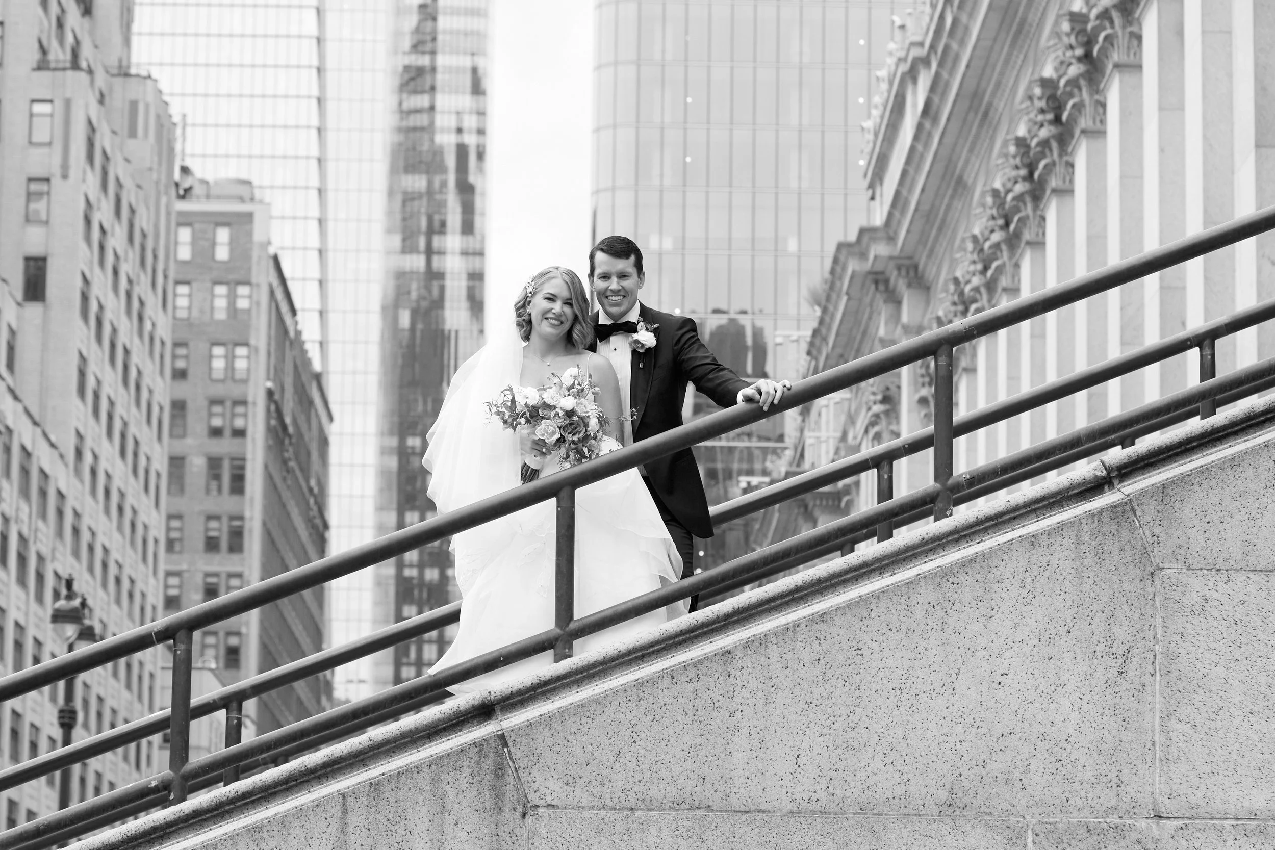 A bride and groom standing on a city bridge, smiling, with tall buildings in the background.