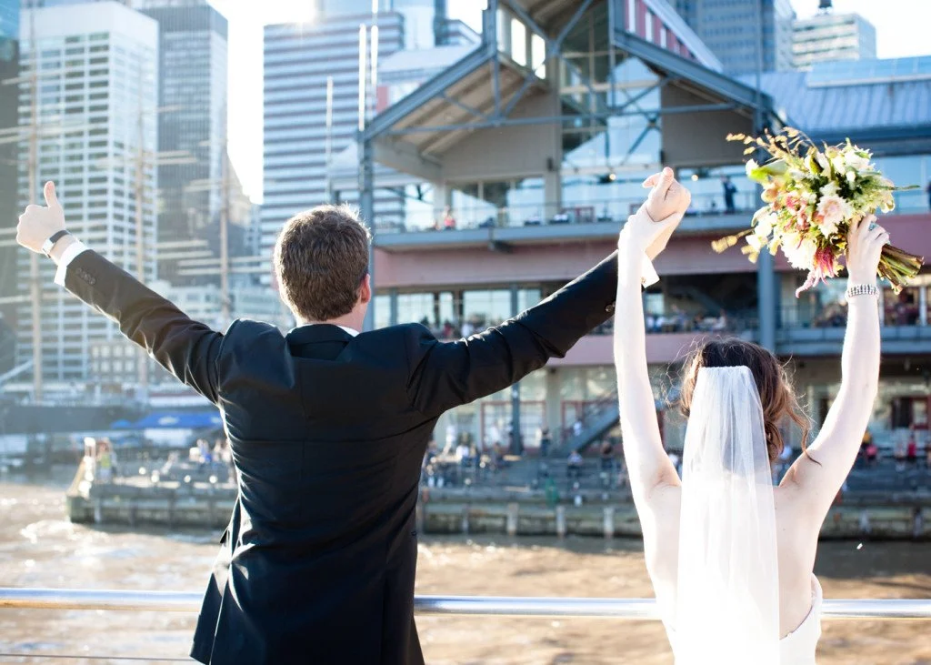 Newlyweds celebrating by a waterfront with city buildings in the background, the man in a suit with arms raised and the bride holding a bouquet with both arms raised.