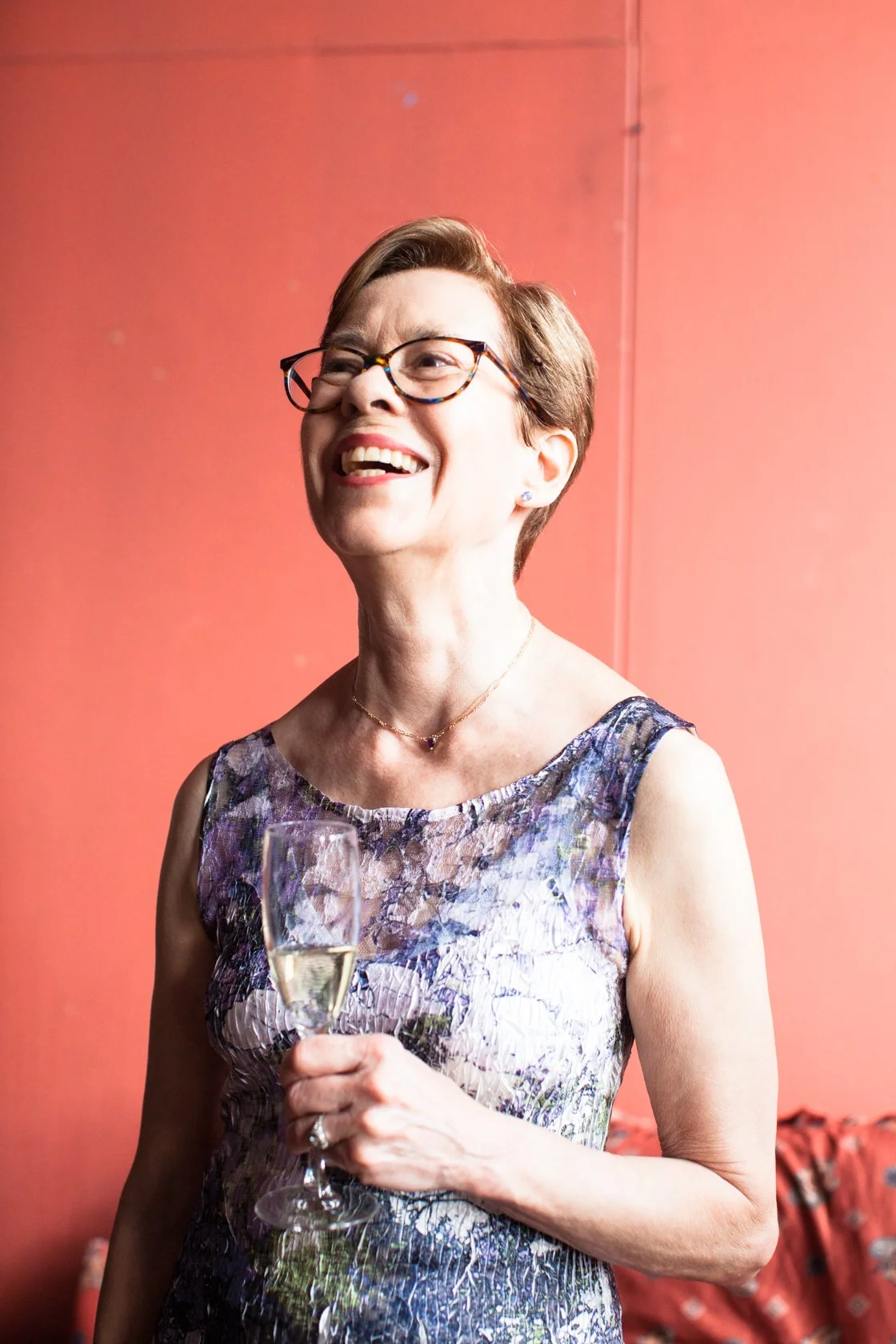 A smiling woman with short hair, glasses, and earrings, wearing a sleeveless floral dress, holding a glass of champagne, standing against a coral-colored background.