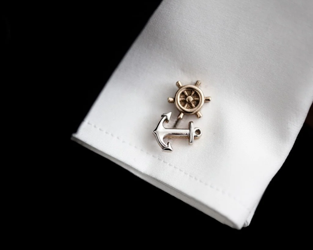 Silver anchor and gold ship's wheel cufflinks on white fabric, against black background.
