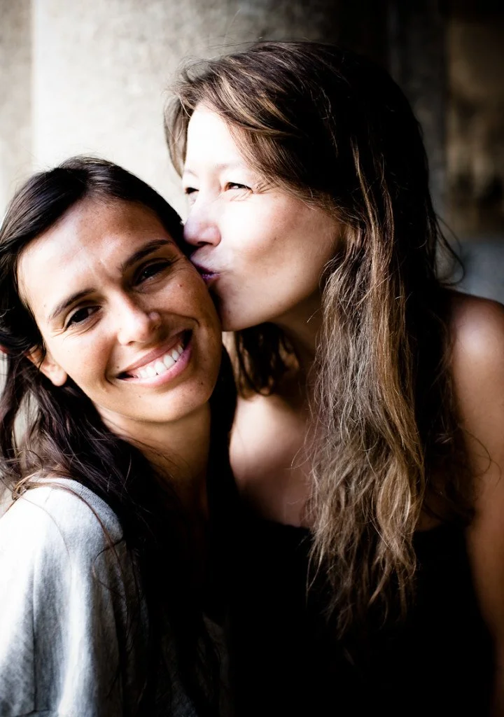 Two women sharing a moment of affection, one smiling at the camera while the other kisses her on the cheek.