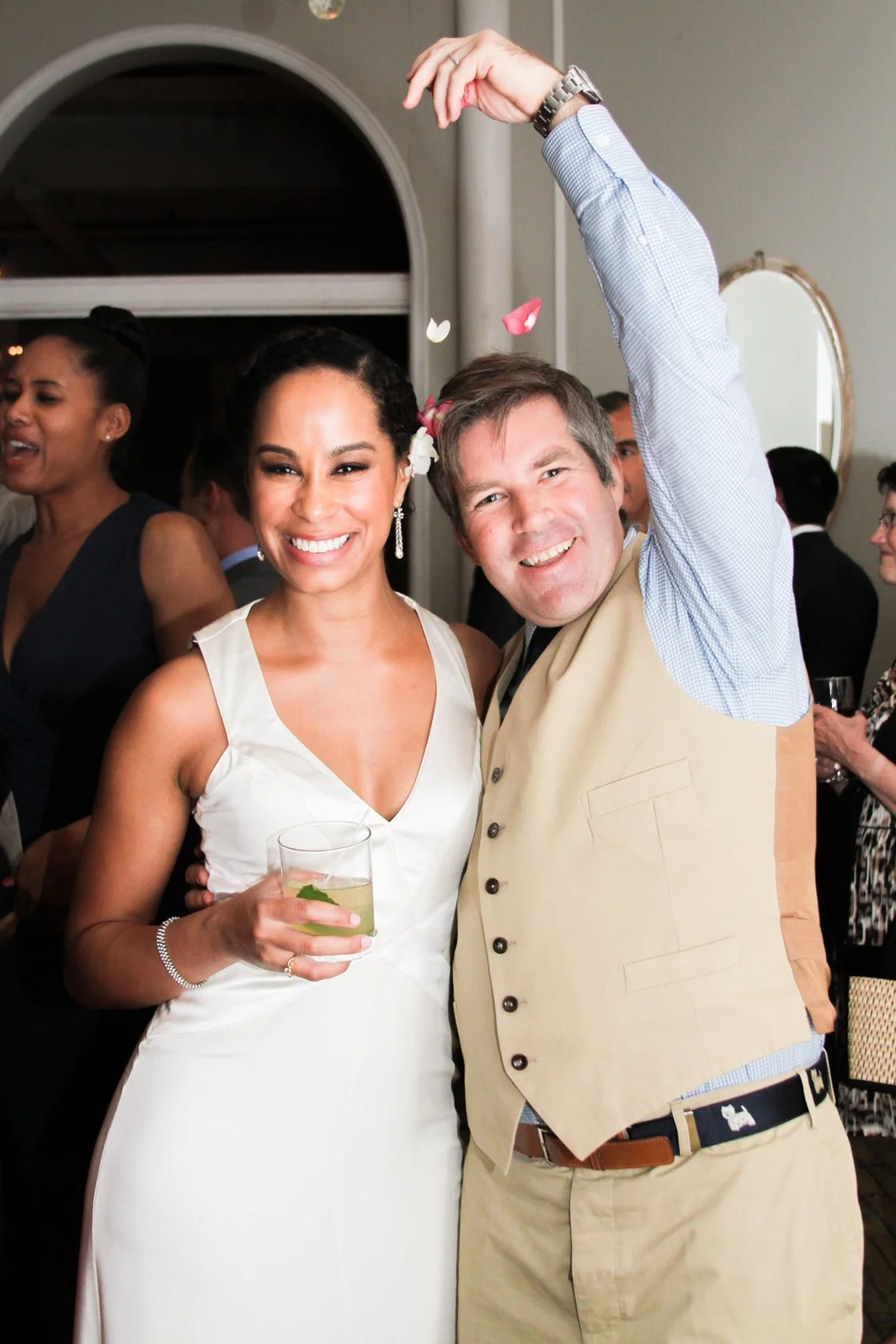 A smiling woman in a white dress holding a drink with mint, standing next to a man in a beige vest and blue shirt at a social event or wedding.