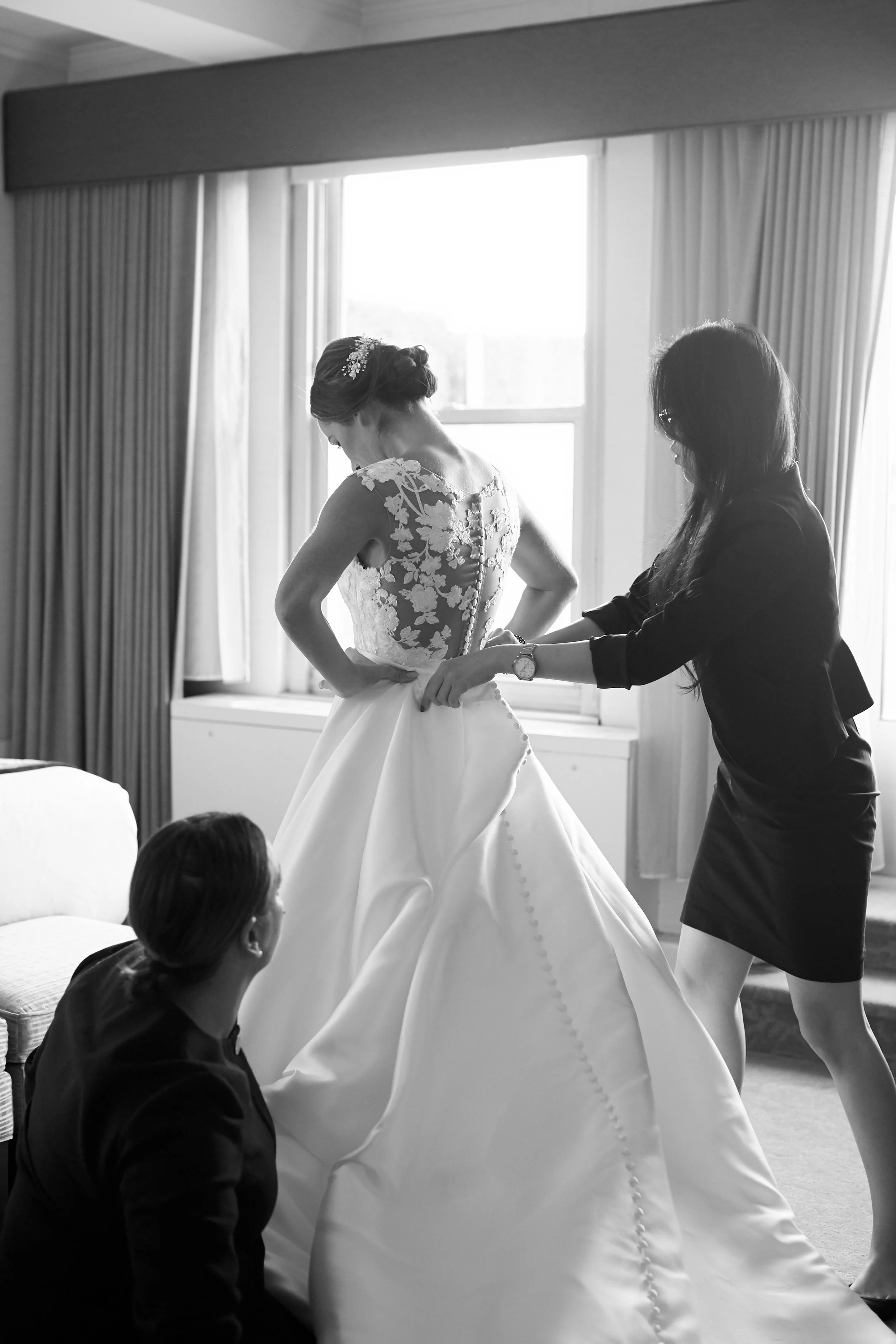 A bride in a wedding dress is being assisted by two women in a room with large windows and curtains.