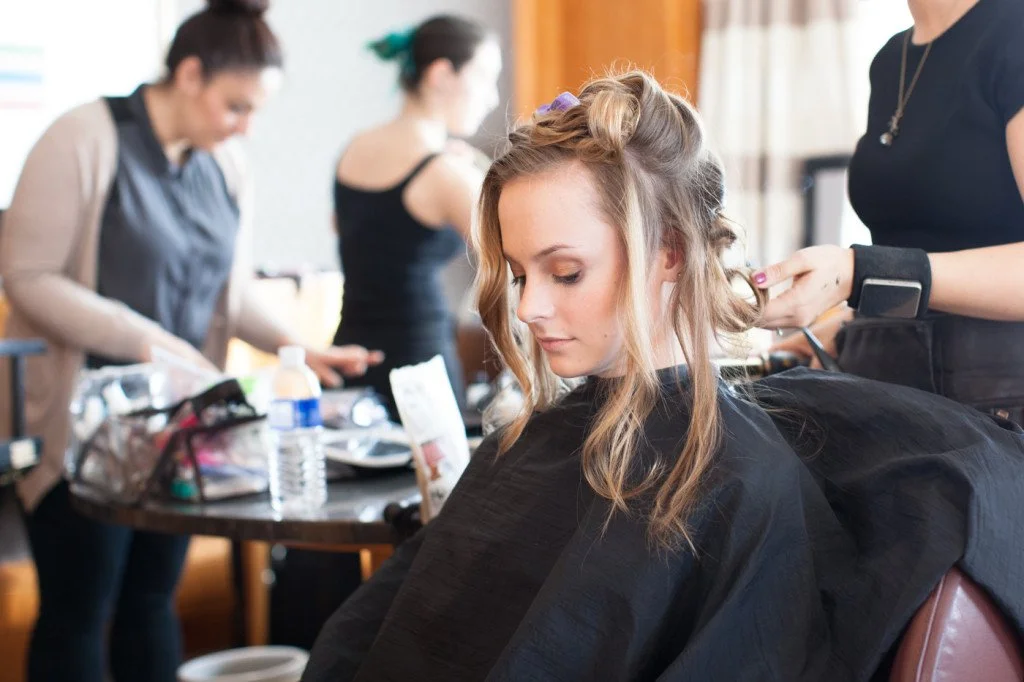 Young woman getting her hair styled by a hairstylist in a salon, with other hairstylists working in the background.