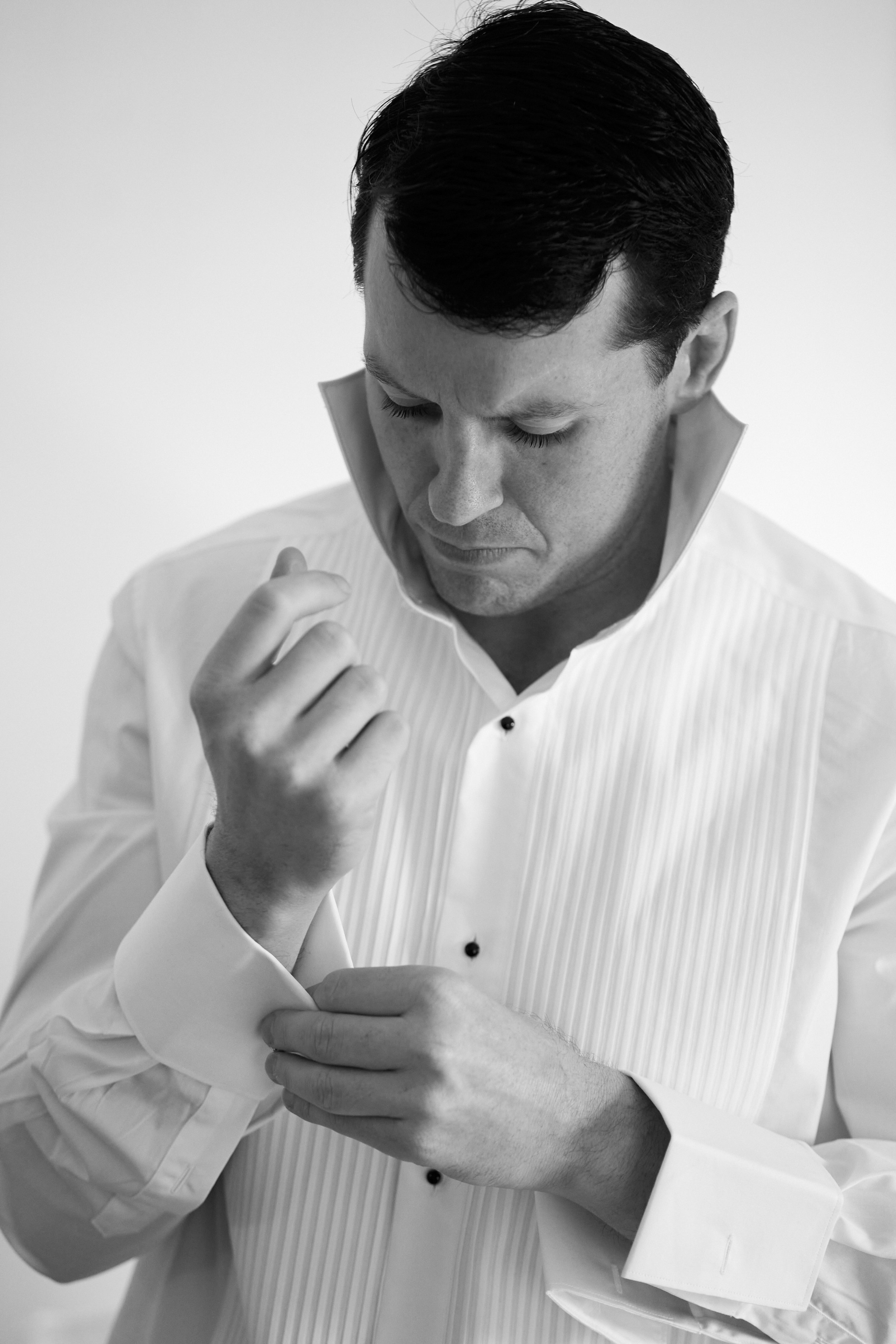 A man with dark hair wearing a white dress shirt with a pleated front, looking down and adjusting his cufflink.