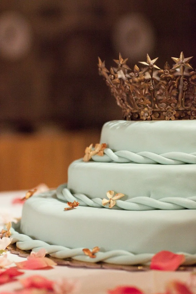 Close-up of a multi-tiered cake with light blue icing, decorated with pink flower petals, small dried flowers, twisted rope-like icing, and a crown on top.