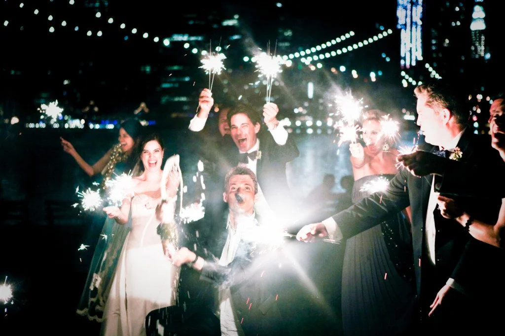 People celebrating at a nighttime event, holding sparklers, with city lights and string lights in the background.