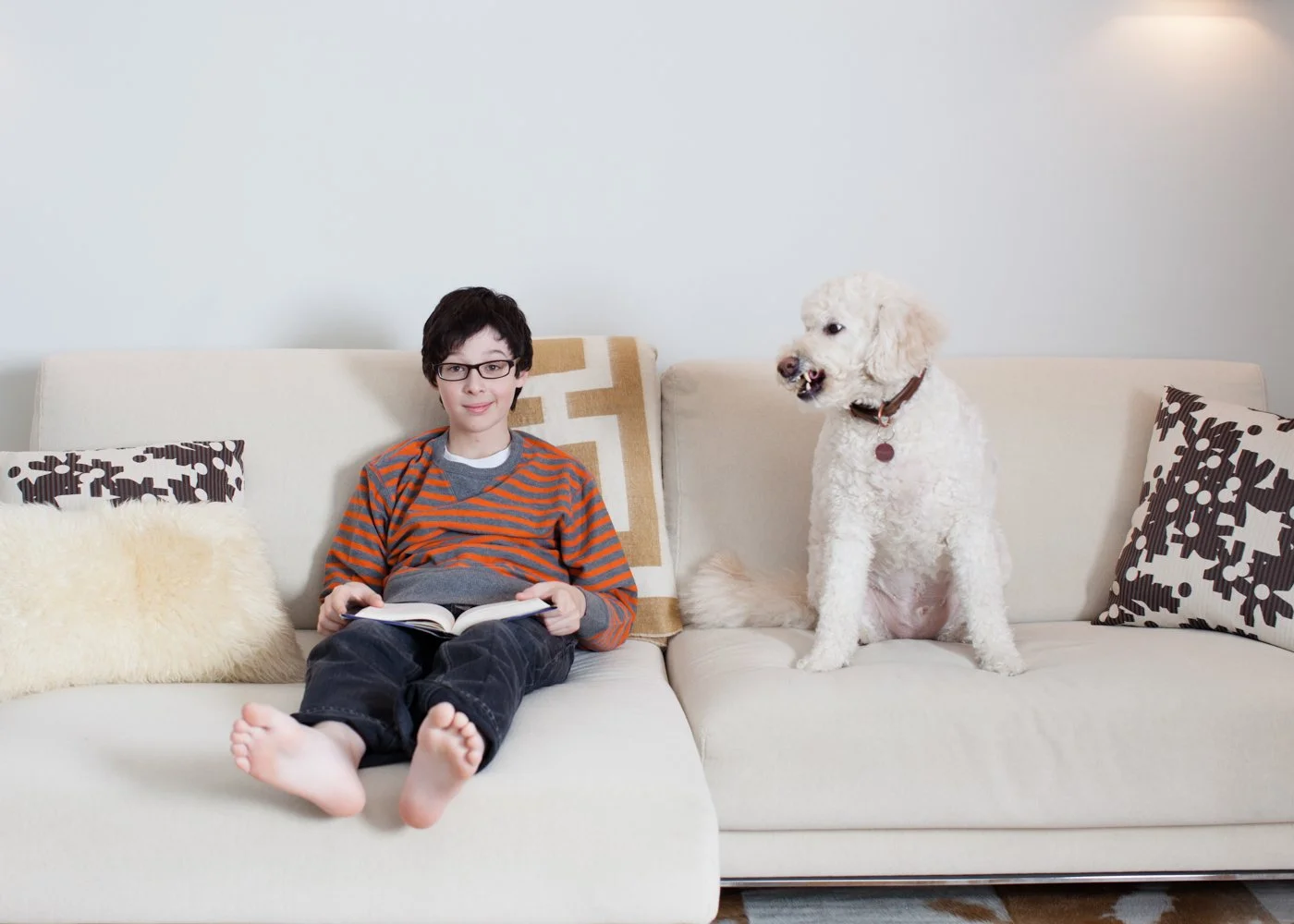 A boy with glasses wearing a striped orange and gray shirt, sitting on a cream-colored couch with a book, next to a large white curly-haired dog sitting on the same couch, against a plain white wall.