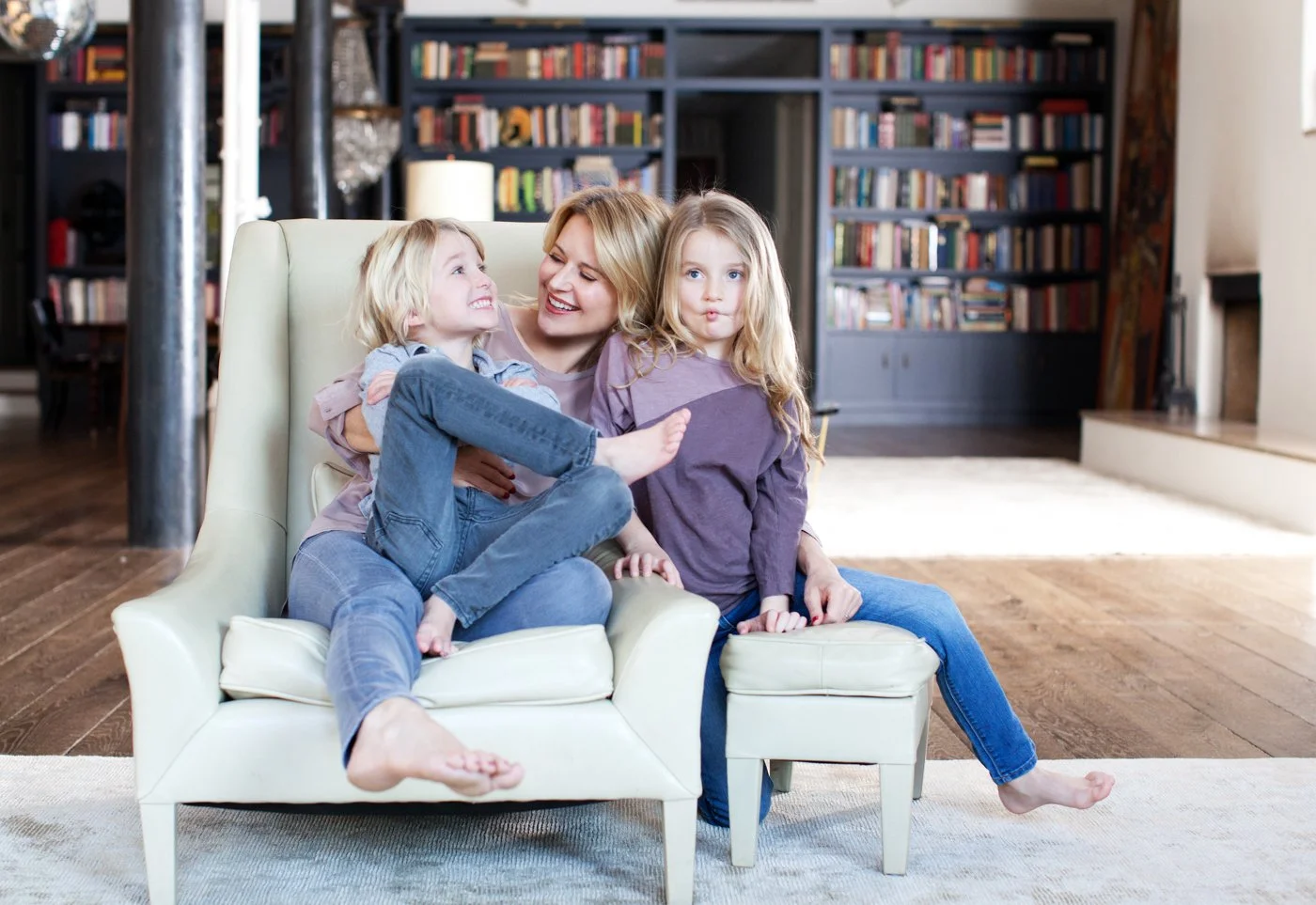 A woman with two young girls sitting on a sofa laughing and playing together in a cozy living room with bookshelves in the background.
