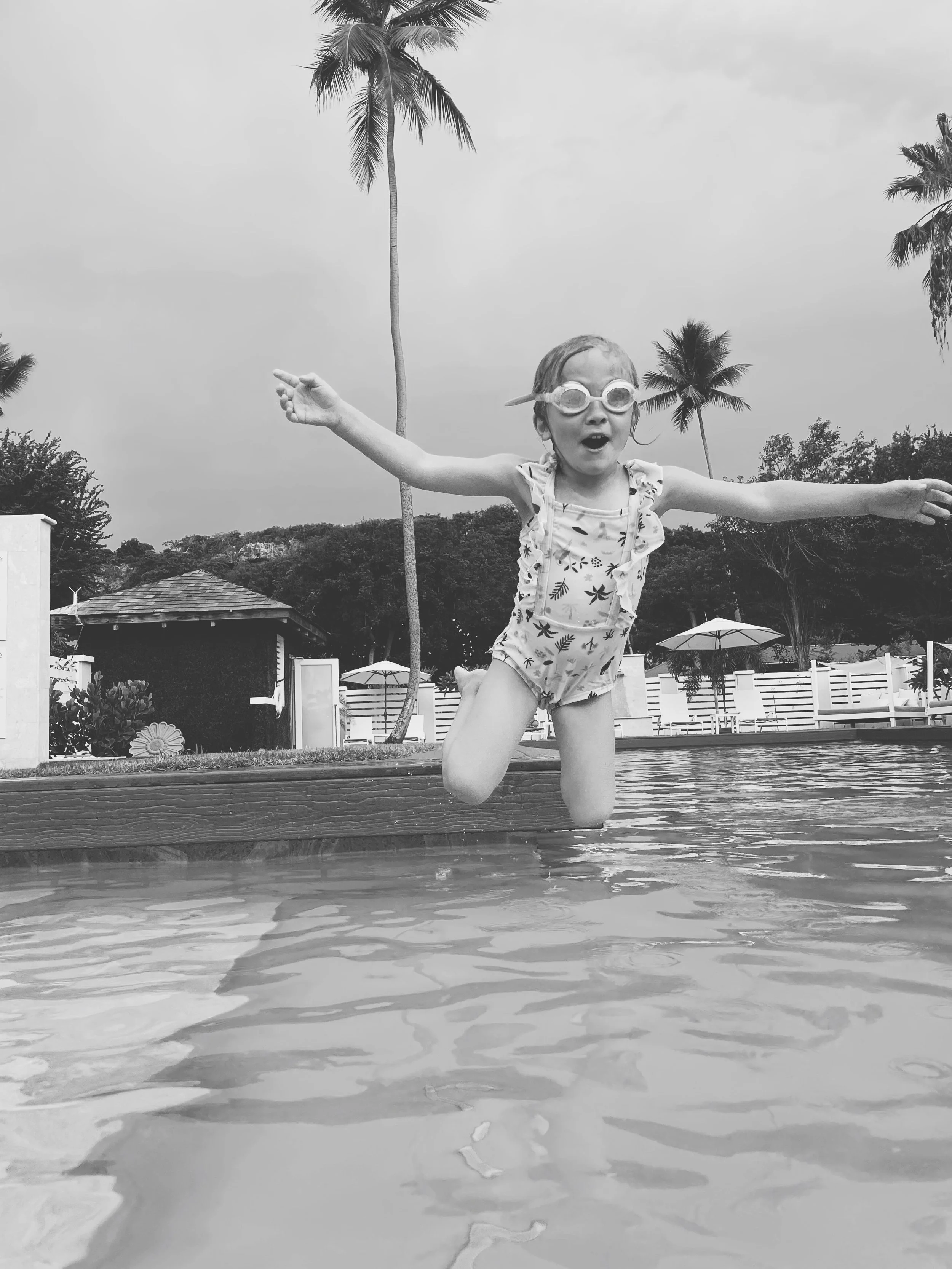 A young girl in a swimsuit and goggles jumps into a swimming pool, with arms outstretched and an excited expression, in a tropical outdoor setting with palm trees and poolside umbrellas.