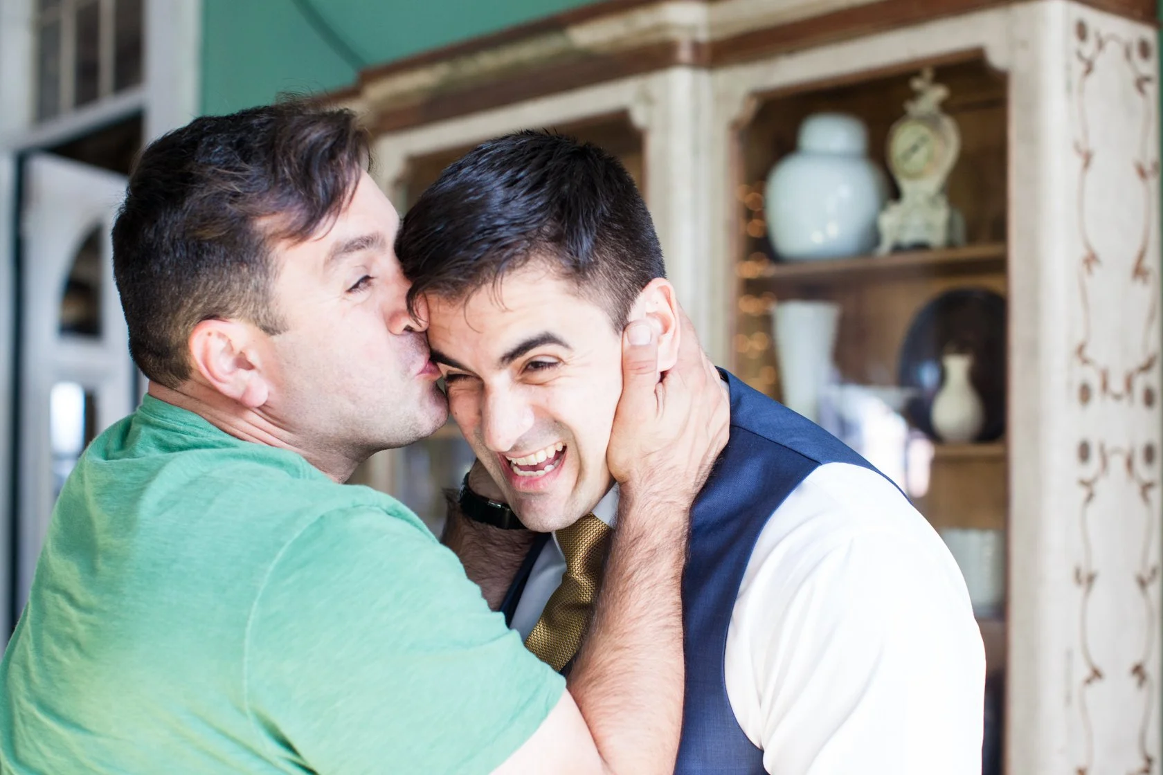 Two men hugging and kissing, with one in a green shirt and the other in a vest and tie, expressing joy and affection.