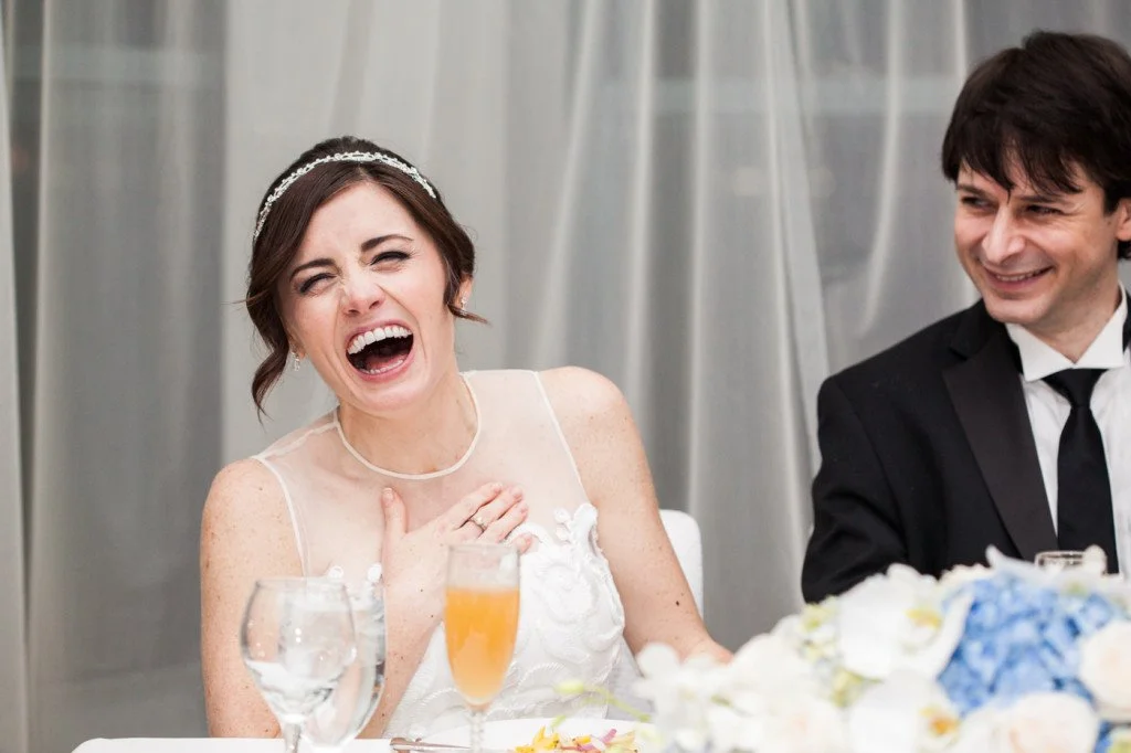 A bride laughing with her hand on her chest, sitting at a table with drinks, beside a groom in a black tuxedo, during a wedding reception.