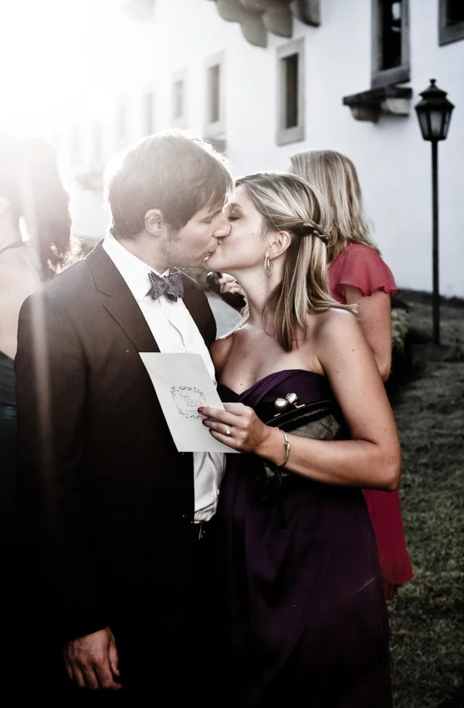 A couple in formal attire kissing outdoors during a wedding or special event, with a guest in a red dress and a traditional white building in the background.