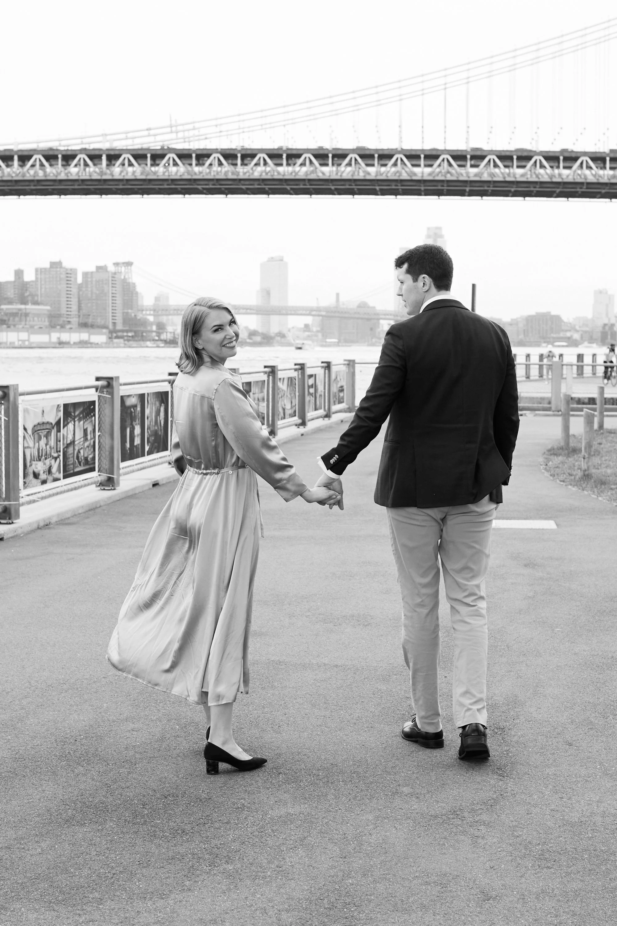 A young couple holding hands and walking along a riverside path in a city, with a bridge and city skyline in the background, in black and white.
