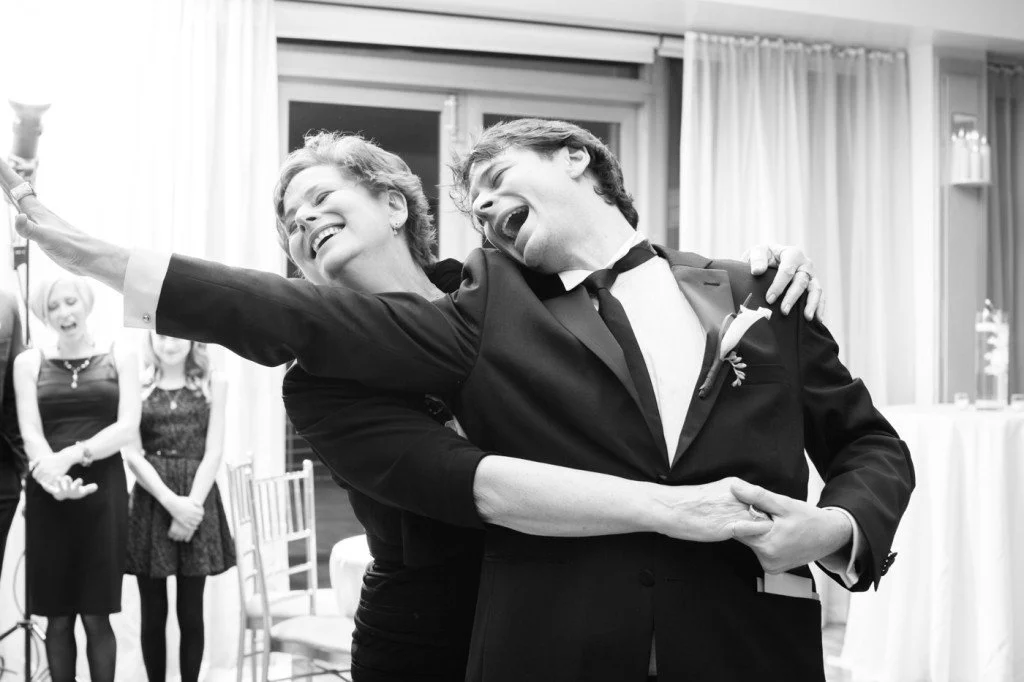 A woman and a man are dancing closely and smiling at a celebration, possibly a wedding, with other guests in the background.