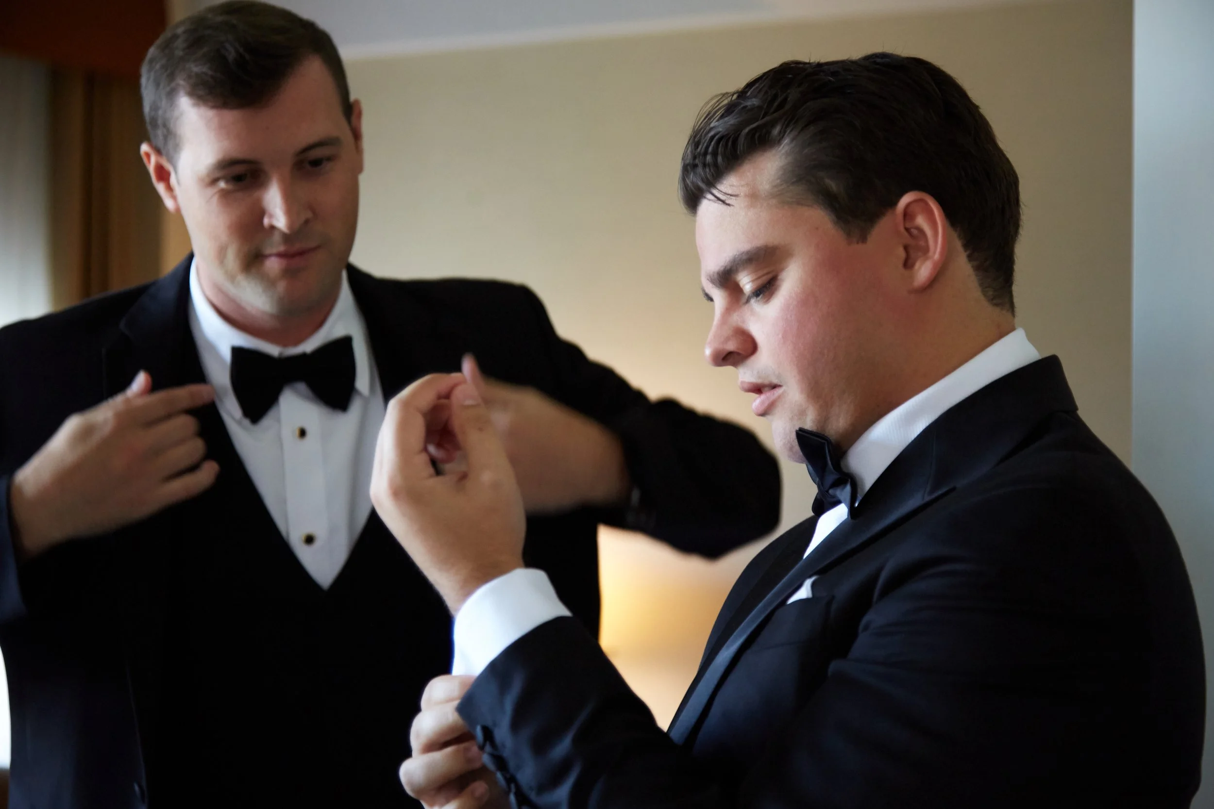 Two men in tuxedos with bow ties getting ready, one helping the other adjust his cufflinks indoors.