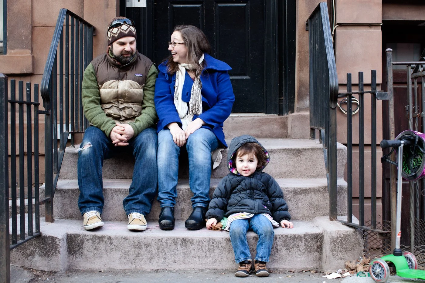 A family of three sitting on the front steps of a brownstone. The father and mother are engaged in conversation, while the young daughter sits in front of them on the lower step, looking at the camera. The family is dressed warmly for cold weather.