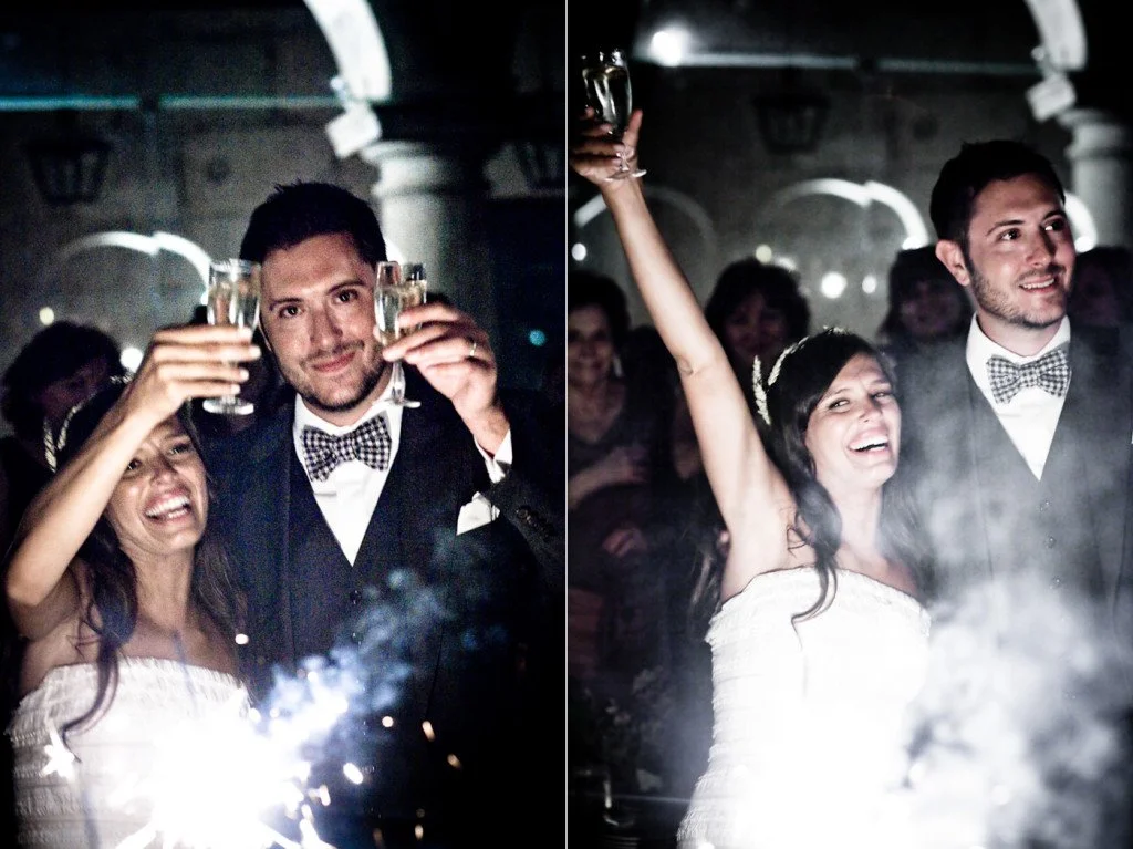 Couple celebrating at a wedding reception, holding champagne glasses and smiling, with sparklers in the foreground and fireworks in the background.