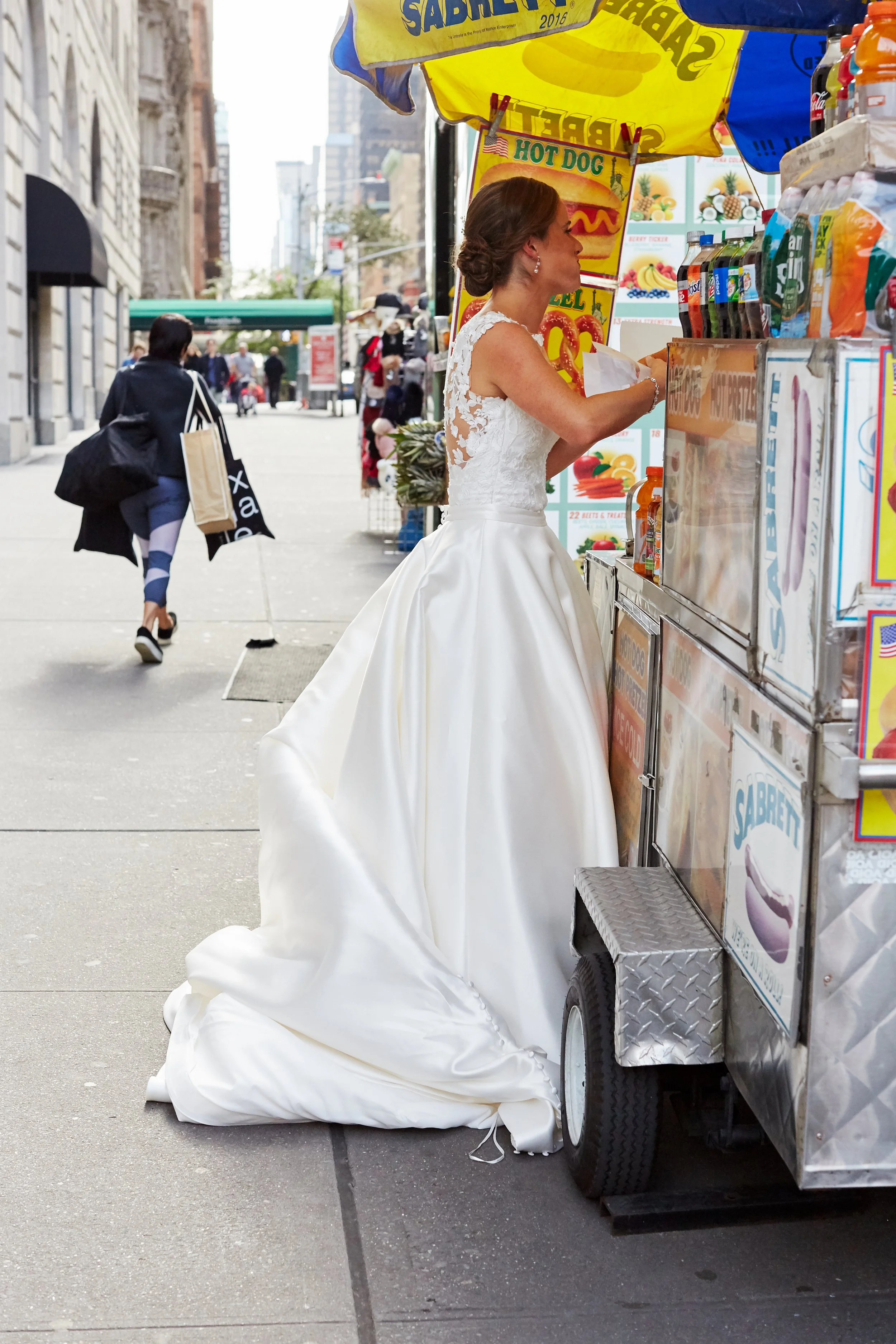 A woman in a wedding dress standing at a street hot dog stand.