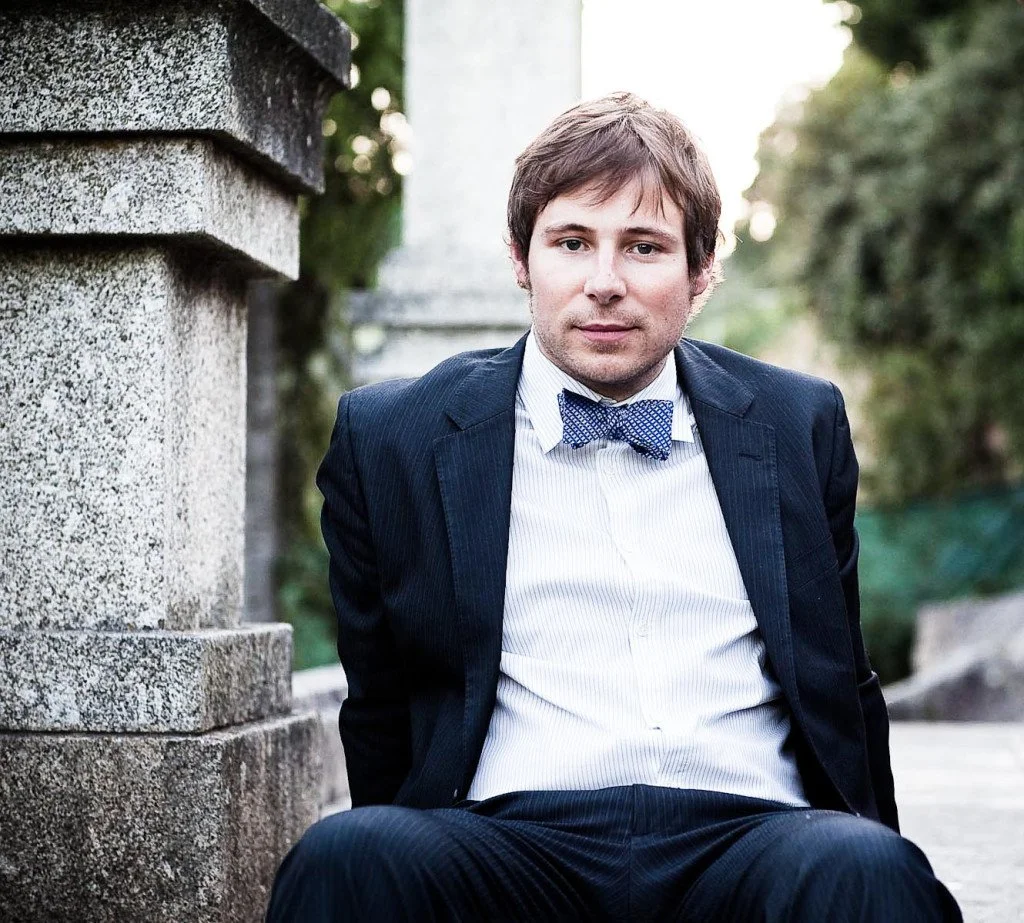 A man in a navy suit and a blue bow tie sitting outdoors near stone pillars and greenery.