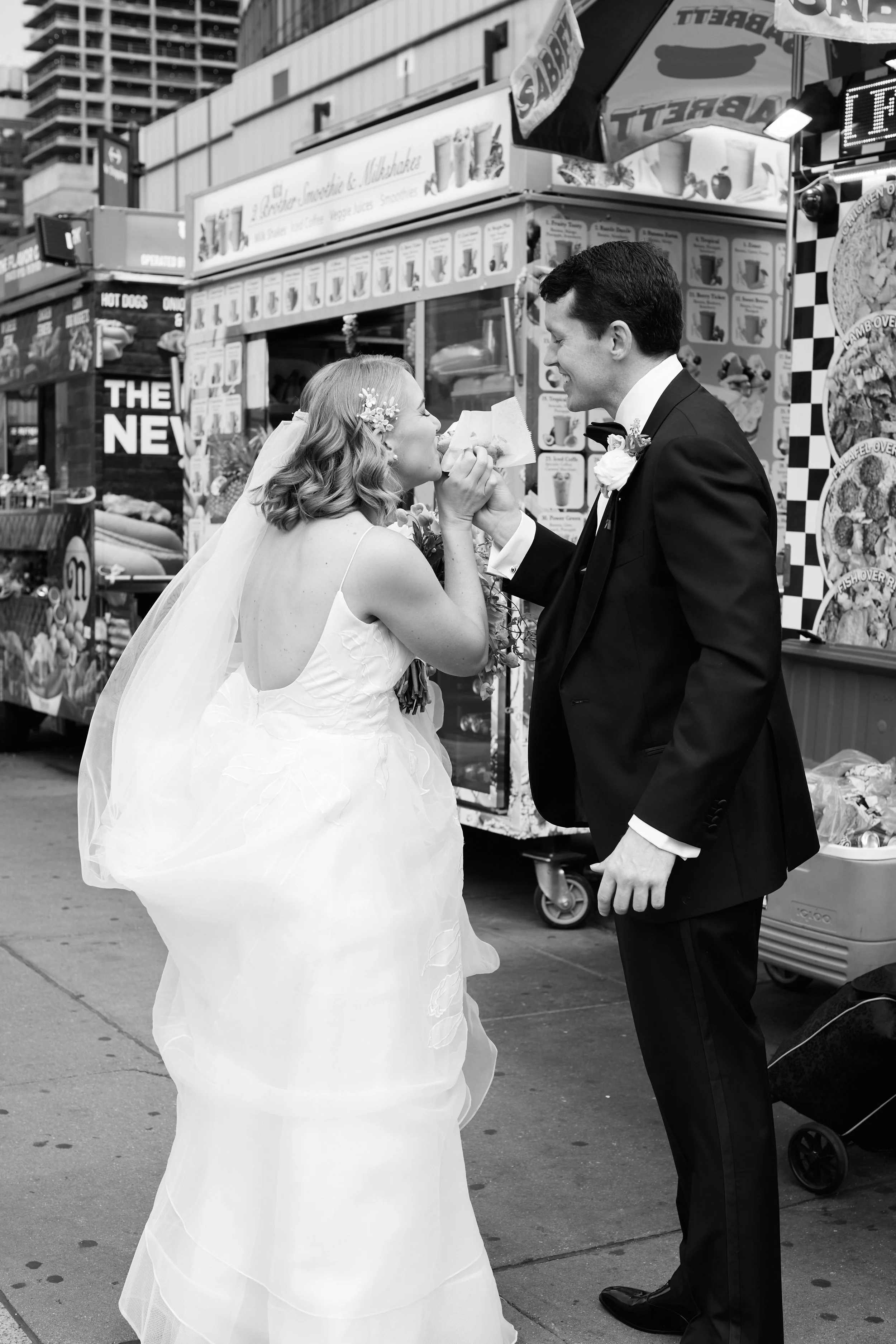 A bride in a wedding dress and veil, and a groom in a tuxedo, sharing a moment on a city street with food stands and advertisements in the background.
