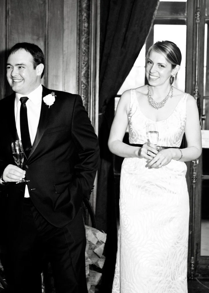 A black and white photo of a man and woman at a formal event. The man is in a tuxedo with a boutonnière, holding a champagne flute. The woman is in a sleeveless white gown, wearing a necklace, and holding a champagne glass, smiling.