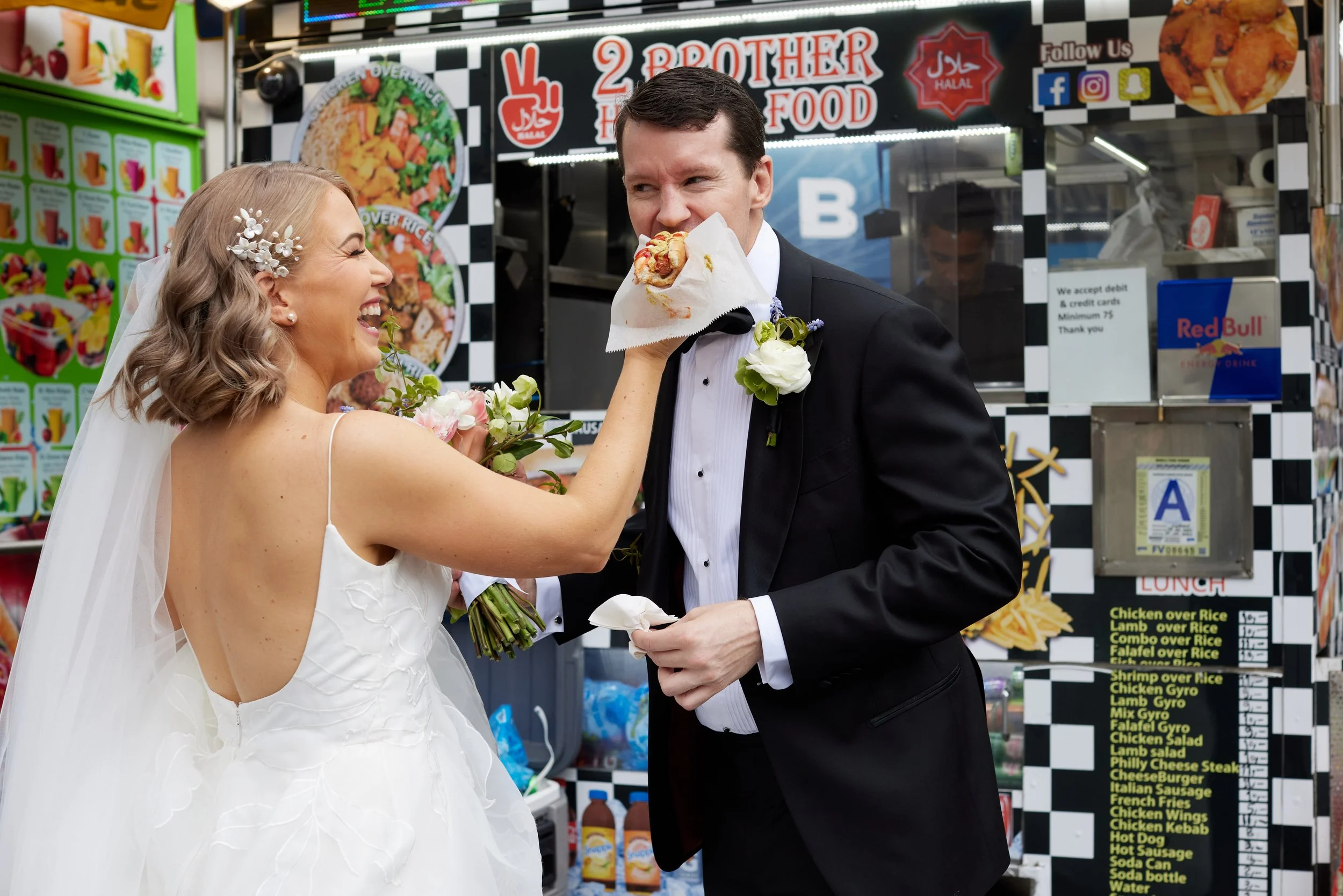A bride playfully tugging a slice of pizza from a groom's mouth inside a fast food restaurant. The bride is wearing a white wedding dress with a veil and floral hairpiece, holding a flower bouquet. The groom is in a black tuxedo with a white shirt an