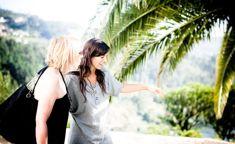 Two women standing outdoors near a palm tree, smiling and pointing at something in the distance.