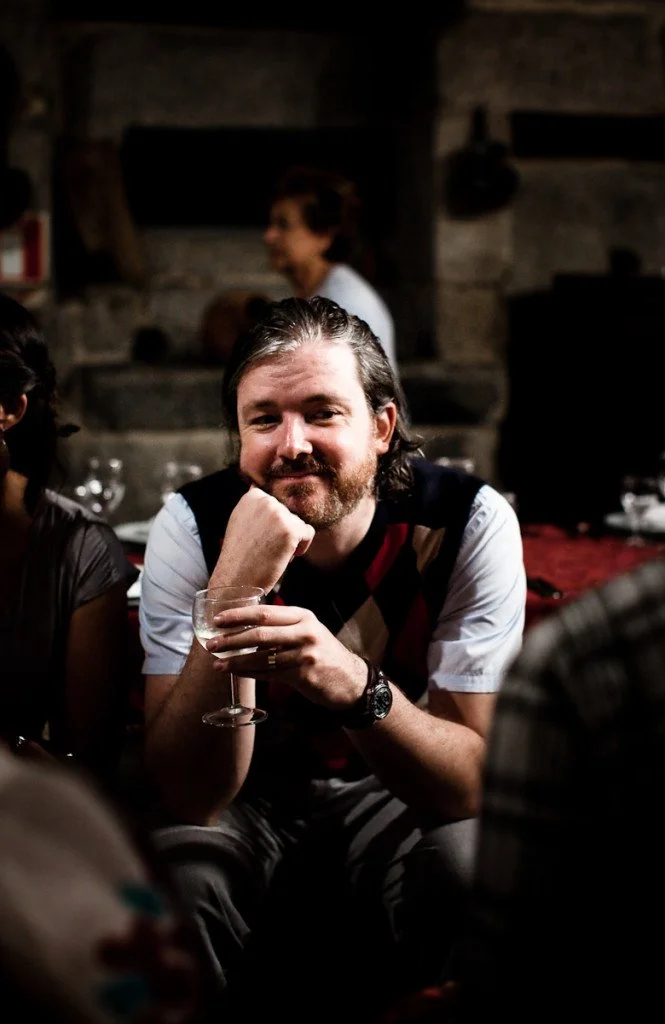 A man with long hair and a beard sitting in a dimly lit room, smiling at the camera, holding a glass of drink, with people and a stone fireplace in the background.
