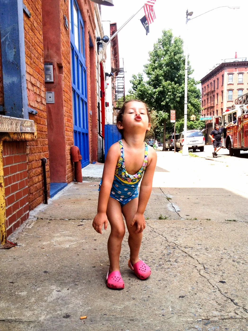 A young girl in a colorful swimsuit and pink Crocs squatting on a city sidewalk, making a playful expression with her lips puckered. Urban background with brick buildings, trees, and parked cars.