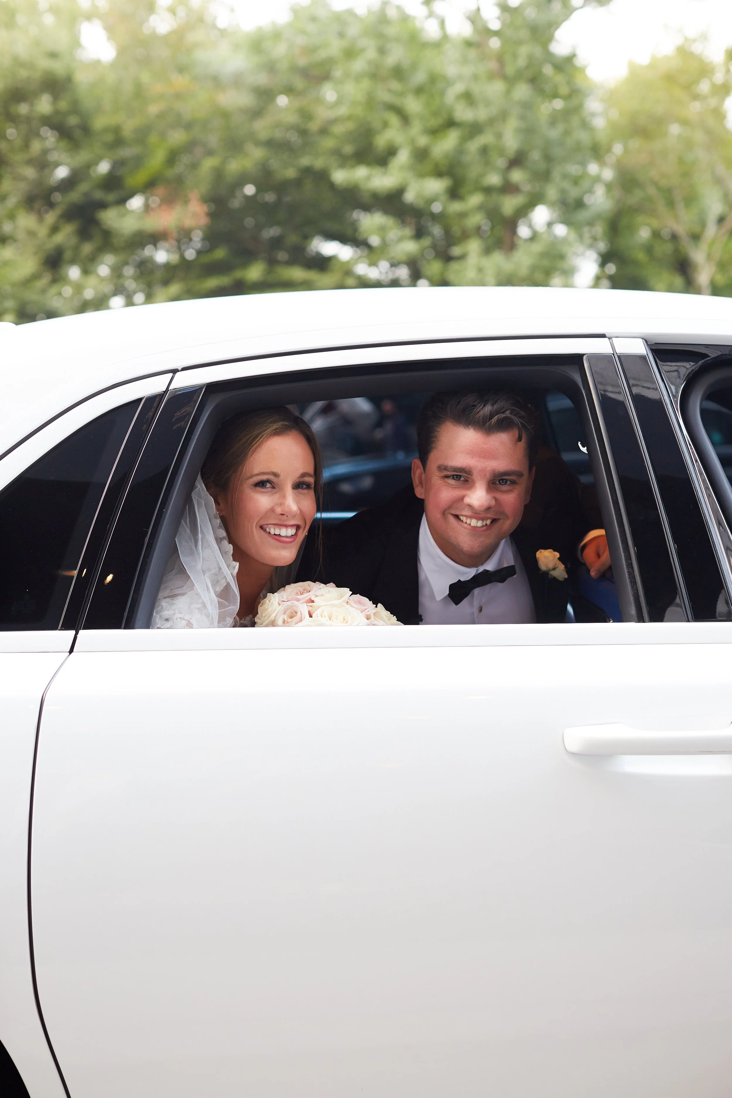 A bride and groom sitting in the back seat of a white car, smiling, with the bride holding a bouquet of white roses, outside with green trees in the background.