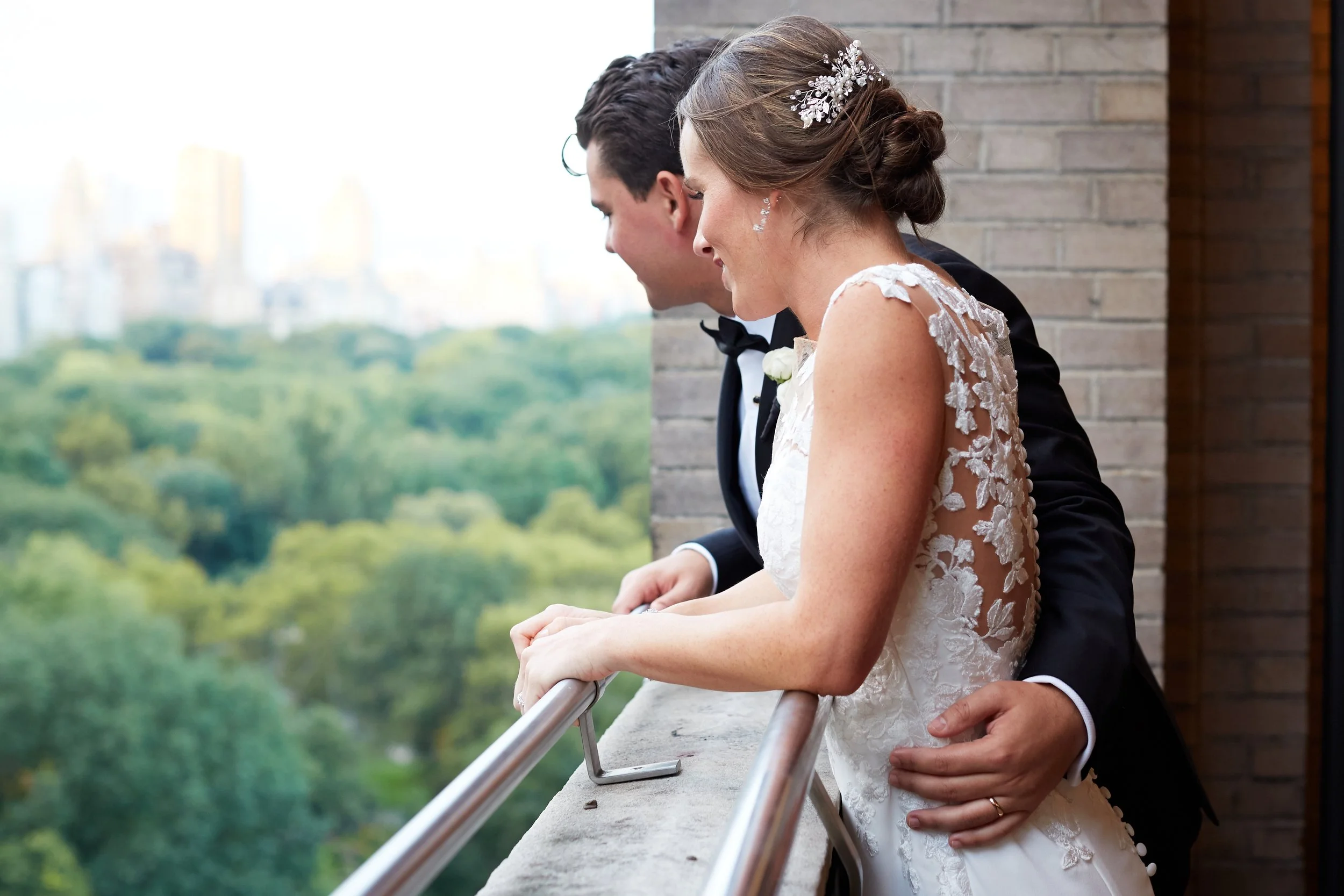 A bride and groom on a balcony, looking out over a city park with tall trees, during their wedding day.