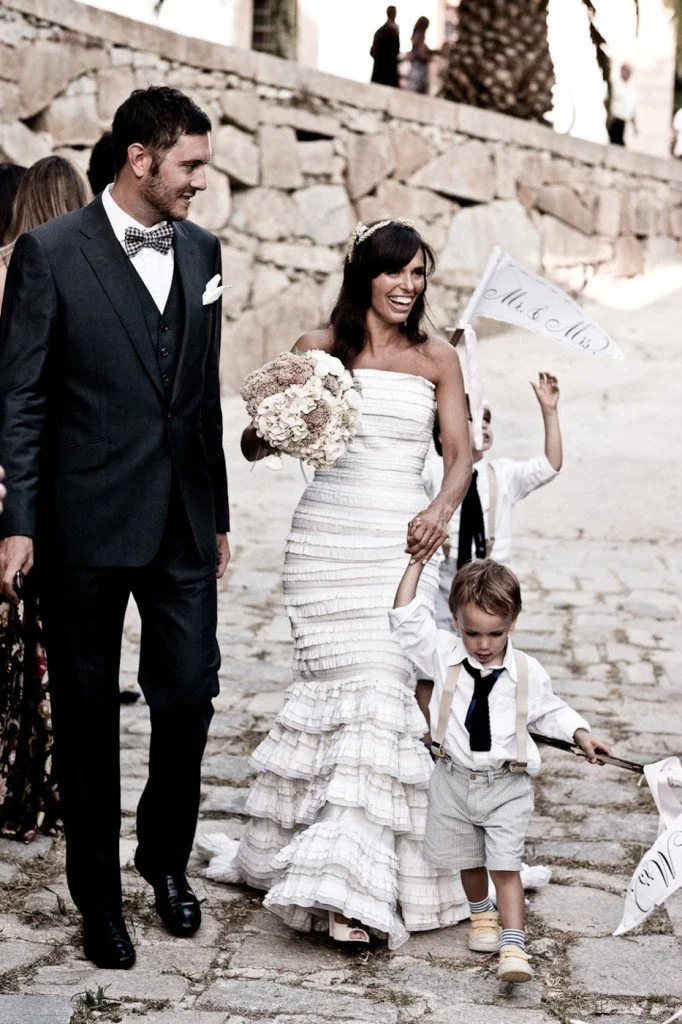 A bride and groom walk hand in hand on a cobblestone street, smiling. The bride wears a white strapless wedding dress and holds a bouquet. The groom wears a dark suit with a bowtie. A young boy in a white shirt, tie, and suspenders walks in front, ho
