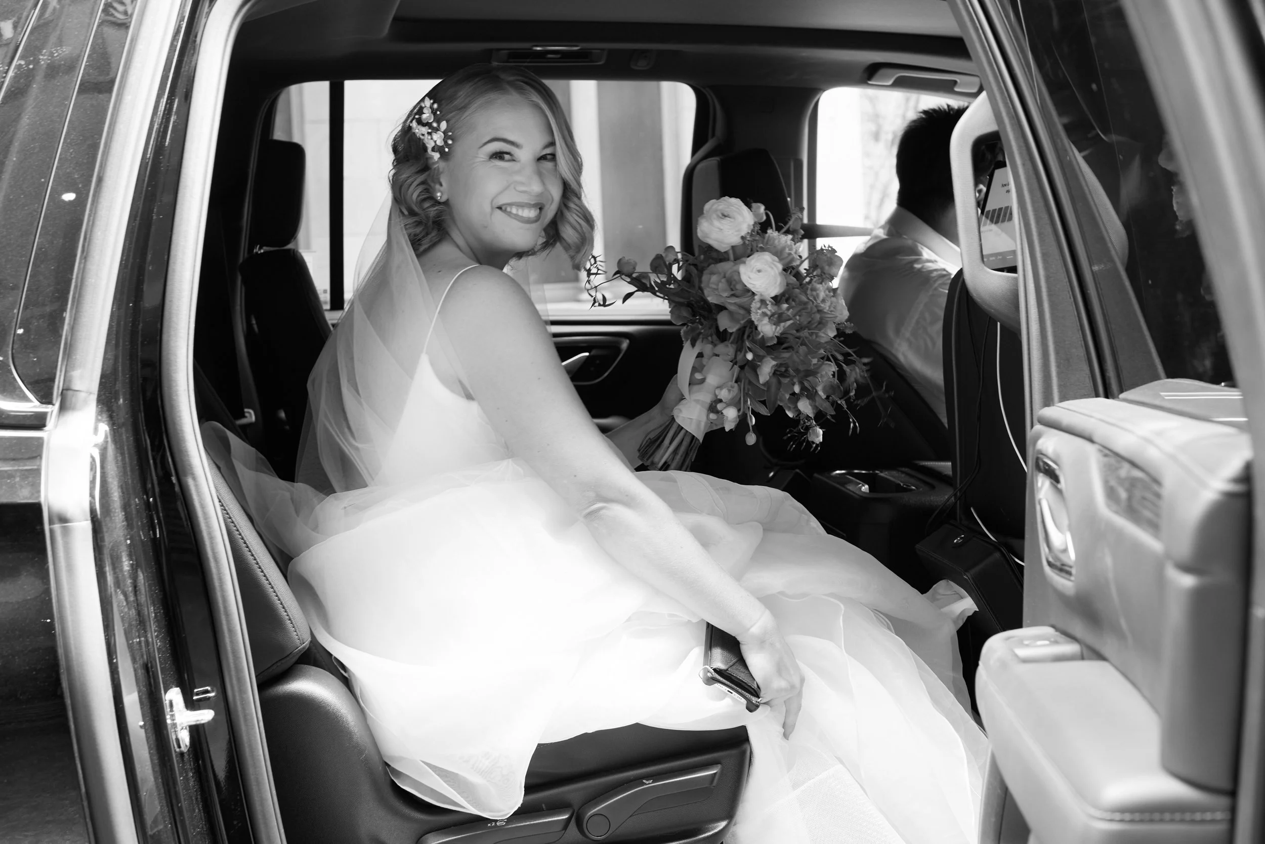 Bride in wedding dress sitting in the back of a vehicle, smiling and holding a bouquet of flowers.