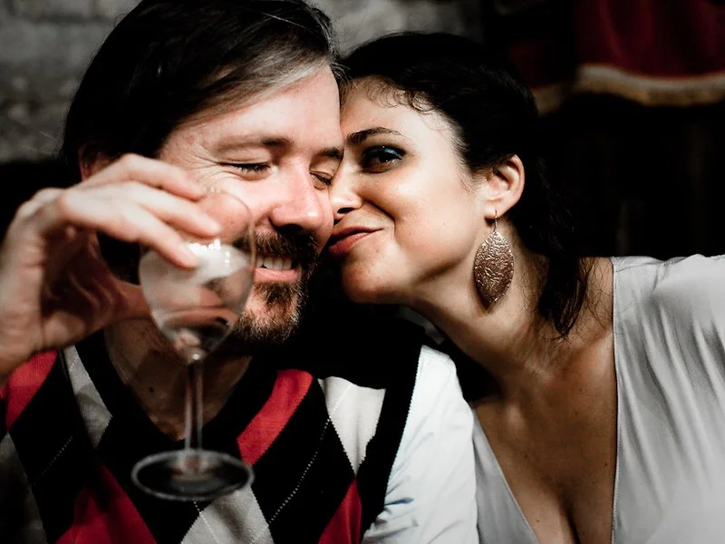 A man with long hair and beard smiling and holding a glass of wine, leaning in close to a woman with dark hair and large earrings, who is smiling and whispering in his ear.