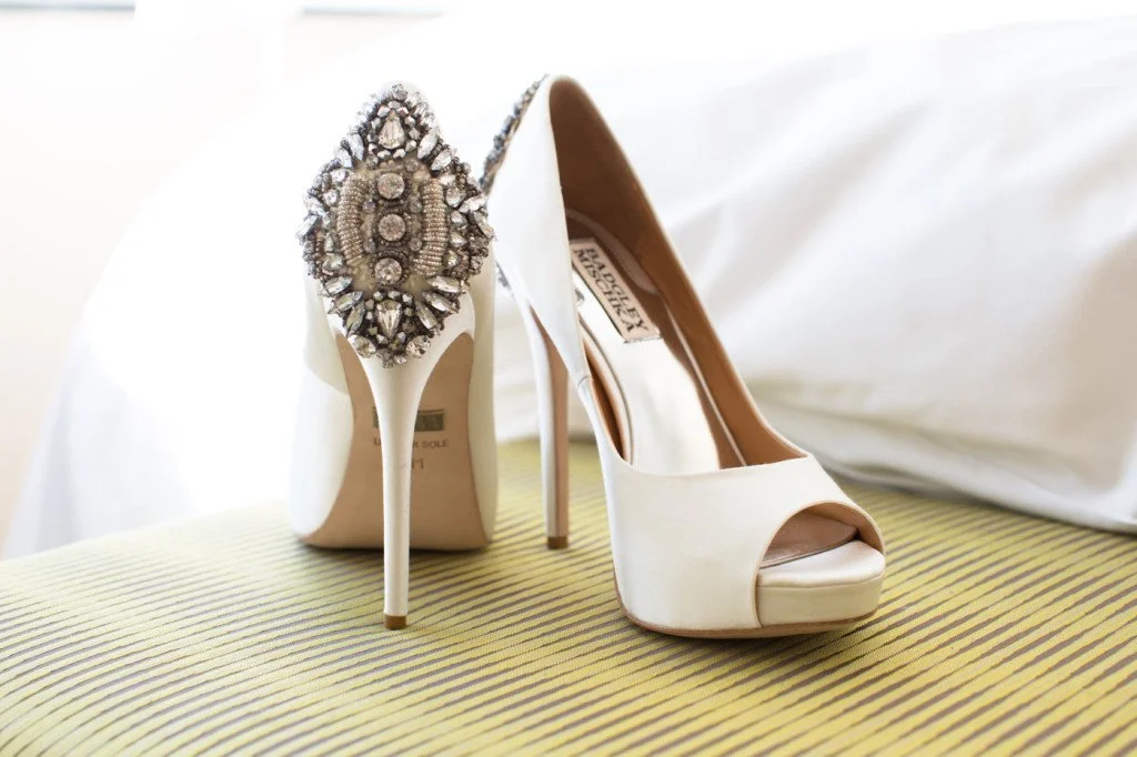 A pair of white high-heeled peep-toe shoes with jeweled embellishments on the back of the heel, placed on a yellow striped surface with a white fabric background.