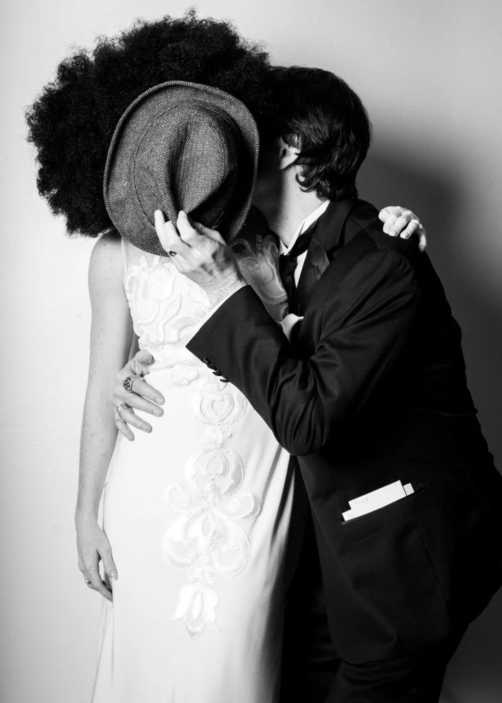 A couple sharing a kiss, with the woman wearing an elegant white dress and the man in a dark suit, holding a hat.