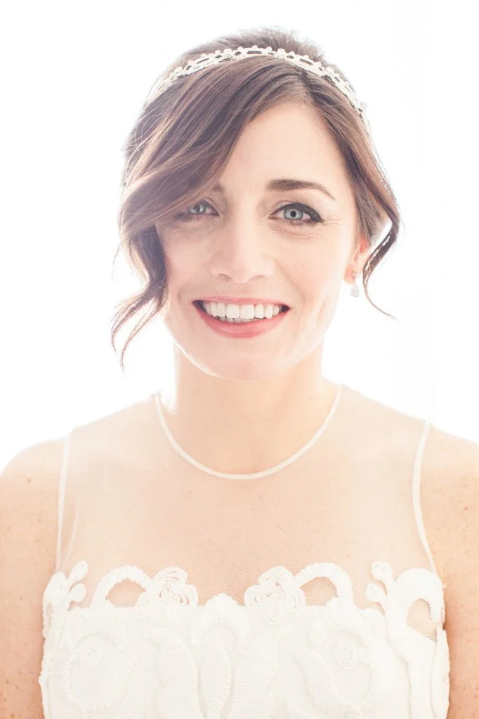 A woman with short brown hair, wearing a white sleeveless dress with lace details, and a pearl headband and necklace, smiling at the camera.