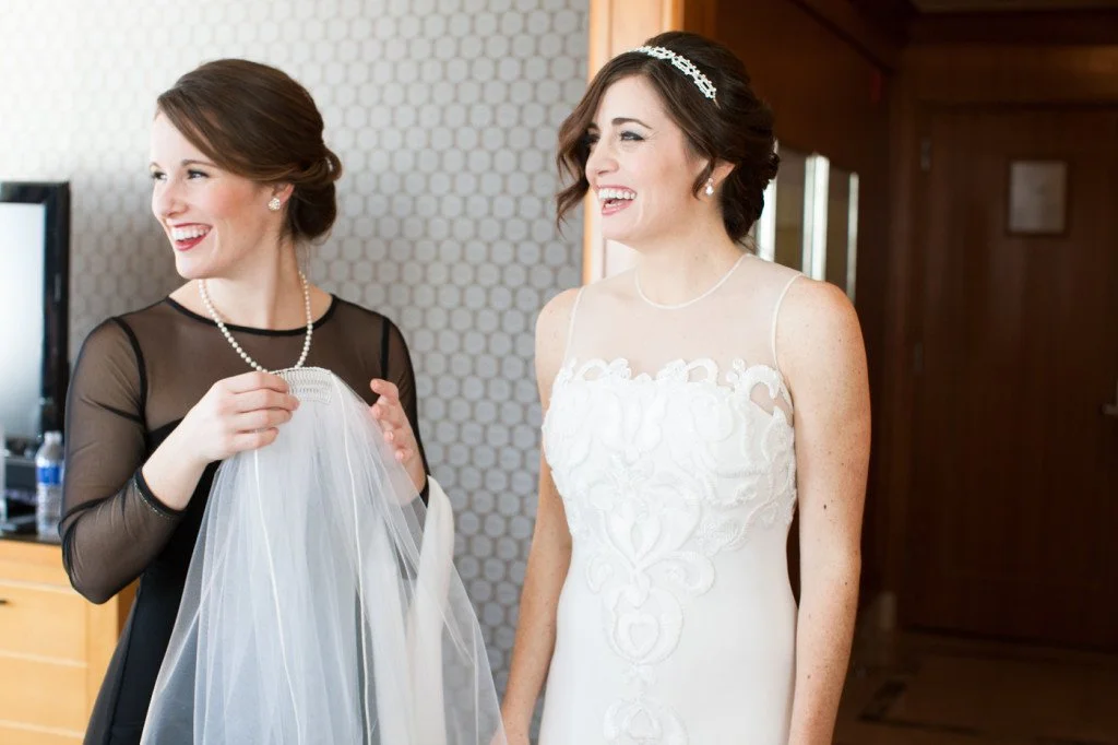 Two women smiling and laughing, dressed in wedding attire, one holding a veil, in a warmly lit room.