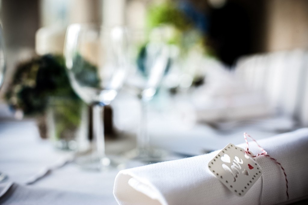 A close-up of a white napkin with a "love" gift tag attached, set on a dining table with wine glasses and blurred background.
