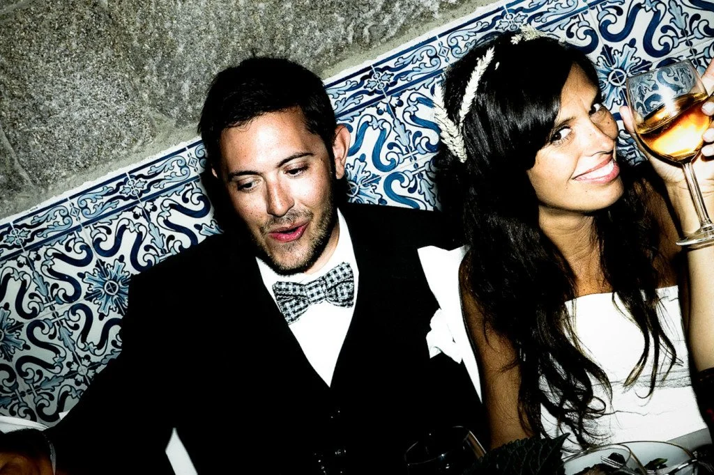 A man and woman sitting together at a table, smiling and toasting with glasses of wine in a restaurant or bar with decorative blue and white tiles on the wall.