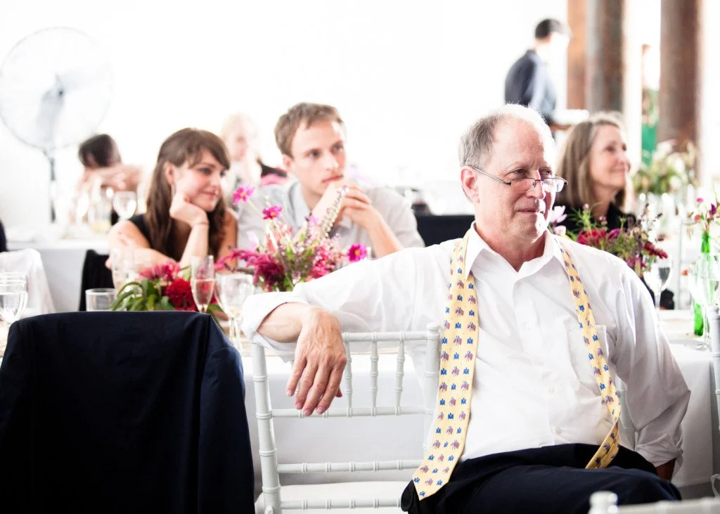 A man with glasses and a white shirt sitting at a banquet table, wearing a colorful tie. Behind him, there are floral centerpieces, glasses, and other guests, including a woman with long hair, a young man, and a woman, attending a formal event or wed