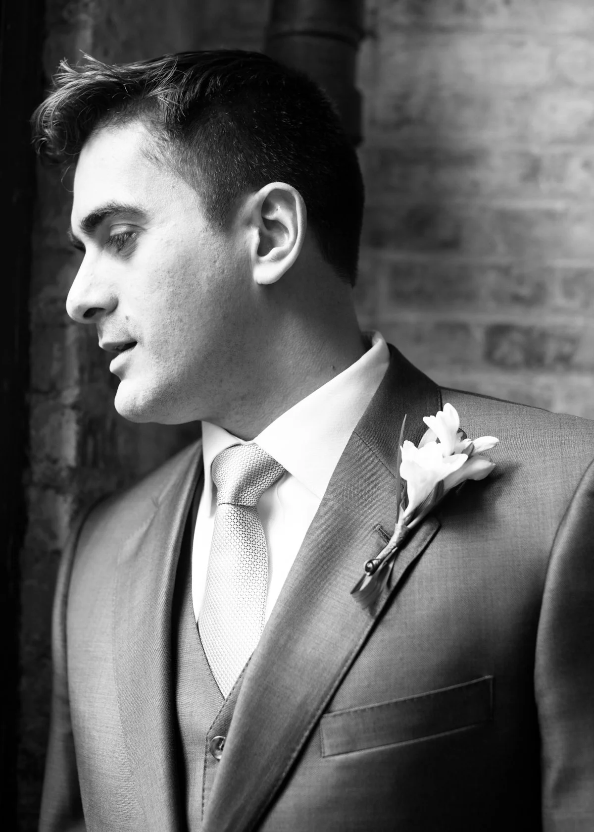 Black and white photo of a man in a suit and tie, with a boutonniere on his lapel, standing against a brick wall.