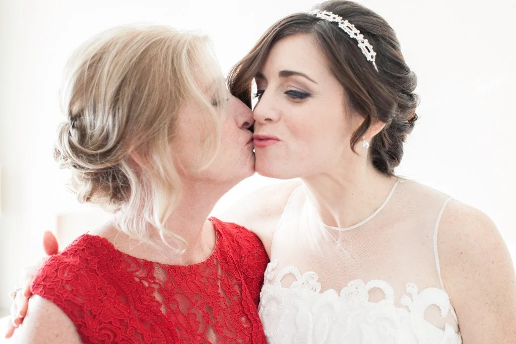A bride in a white wedding dress and a headband receives a kiss on the cheek from an older woman in a red dress.