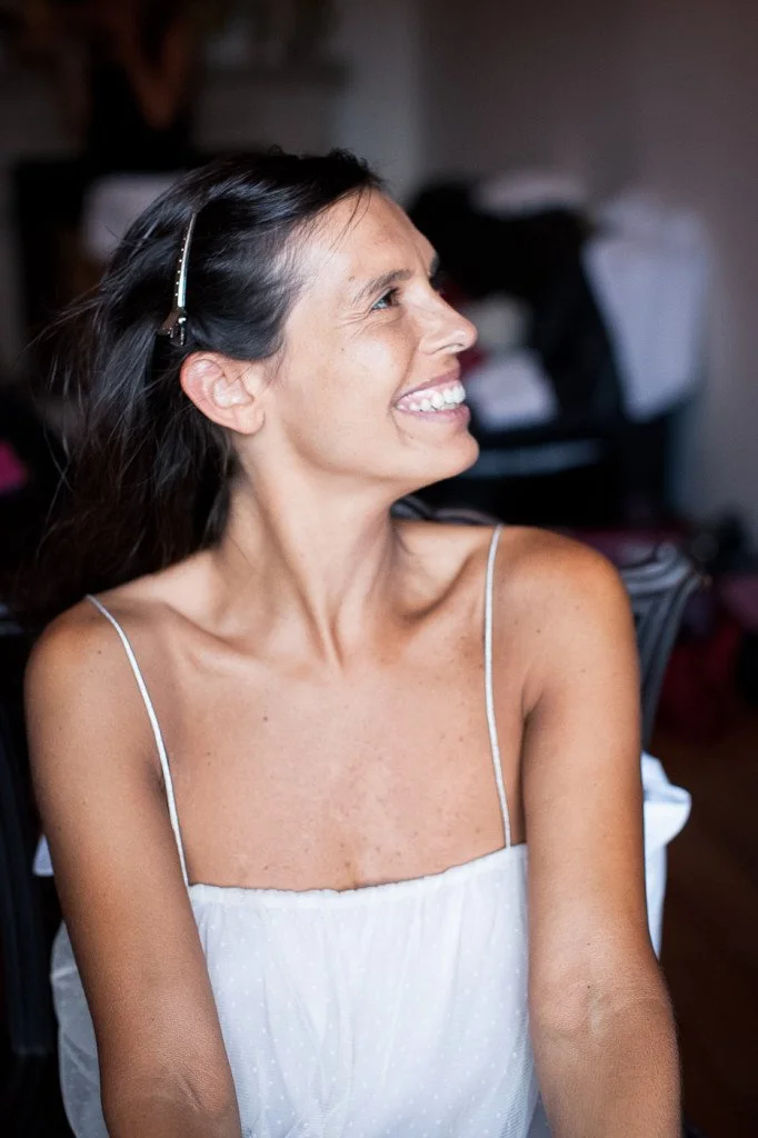 A woman with long dark hair, wearing a white spaghetti strap top, smiling and looking to her right indoors.