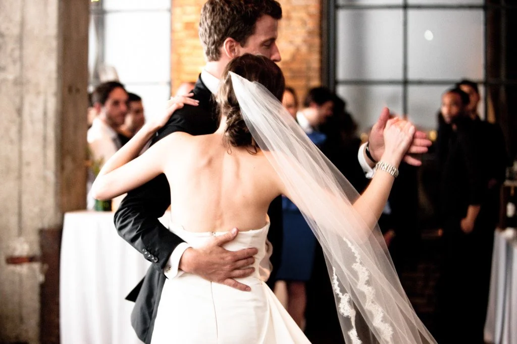 A bride and groom dancing at their wedding reception inside a rustic venue, with guests watching in the background.
