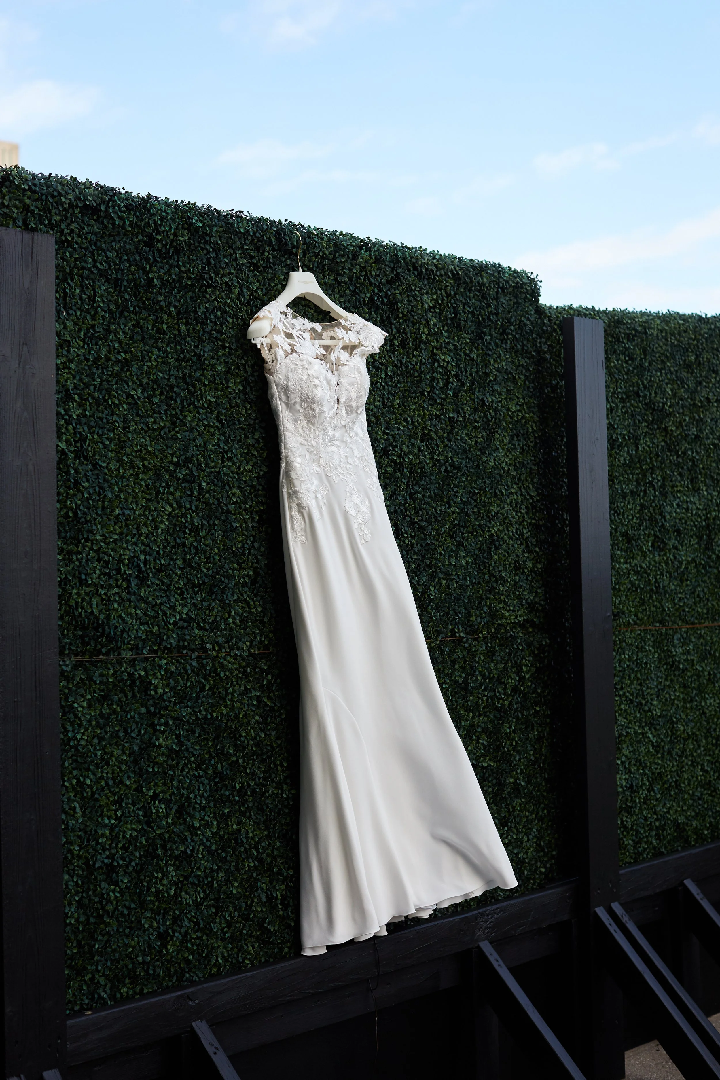 A white wedding dress hangs on a garden hedge against a blue sky.