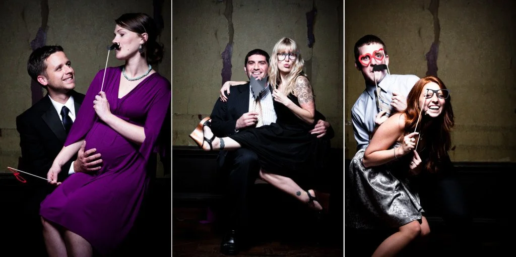 Three groups of people are posing playfully with photo booth props against a dark wall: a pregnant woman in purple with a mustache prop, a man and woman smiling with a sock puppet, and a couple with funny glasses and heart-shaped sunglasses, holding 