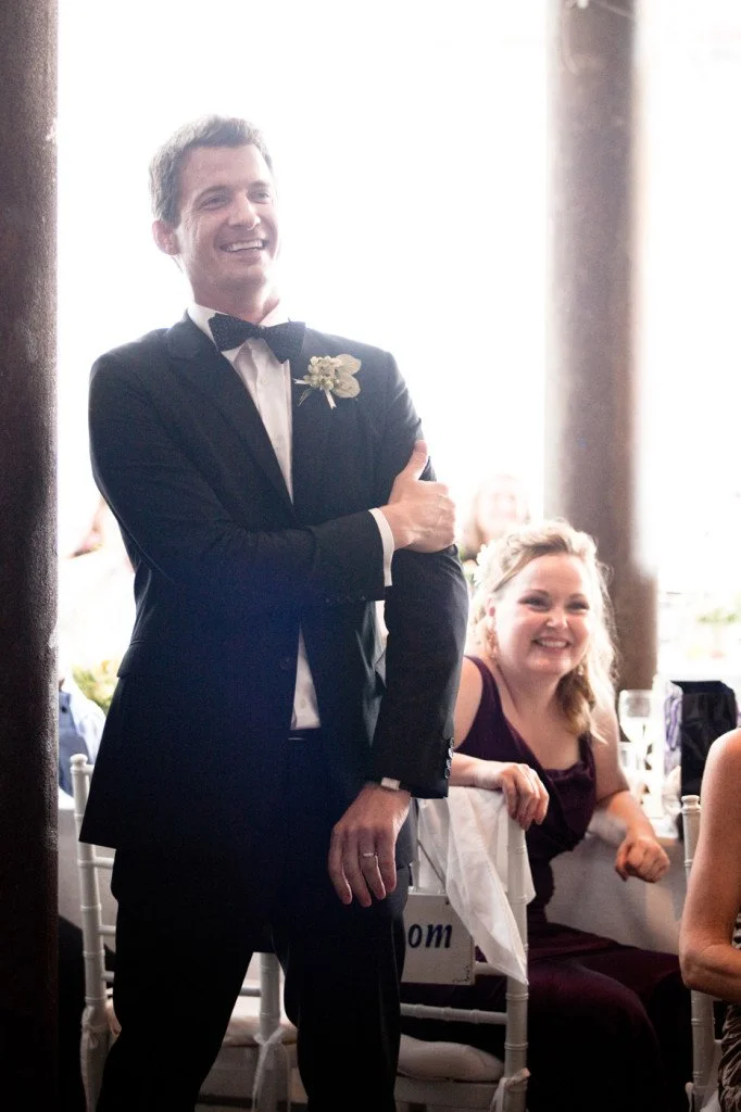 A man in a tuxedo with a boutonnière smiling and standing with his arms crossed, at a formal event, with a woman in a burgundy dress smiling in the background.