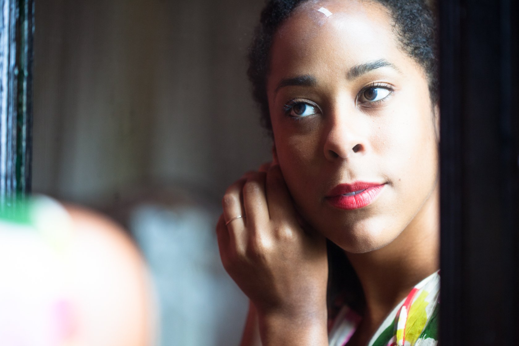 A woman with short, curly hair looking out a window with a pensive expression, resting her chin on her hand.